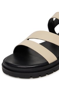 Sandale din piele bej cu două barete încrucișate peste o talpă texturată neagră; au o talpă din cauciuc negru, robustă, cu un model bine definit.