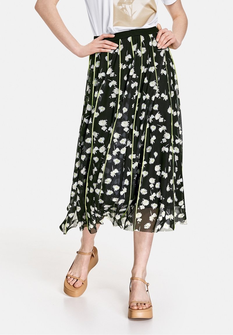 Femme portant une jupe midi noire avec un motif floral blanc et des lignes verticales vertes, associée à des sandales compensées couleur beige.