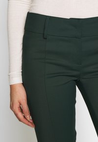 Mörkgröna byxor med en jämn textur, figursydd design och midja med en enkel knappstängning. Sytt detaljarbete längs sidorna.