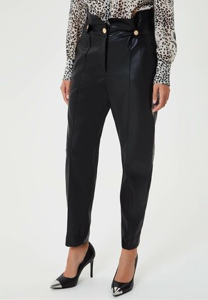 Femme portant un pantalon en cuir noir taille haute avec des boutons dorés, associé à des talons noirs à bouts pointus avec des embouts métalliques et un chemisier imprimé léopard.