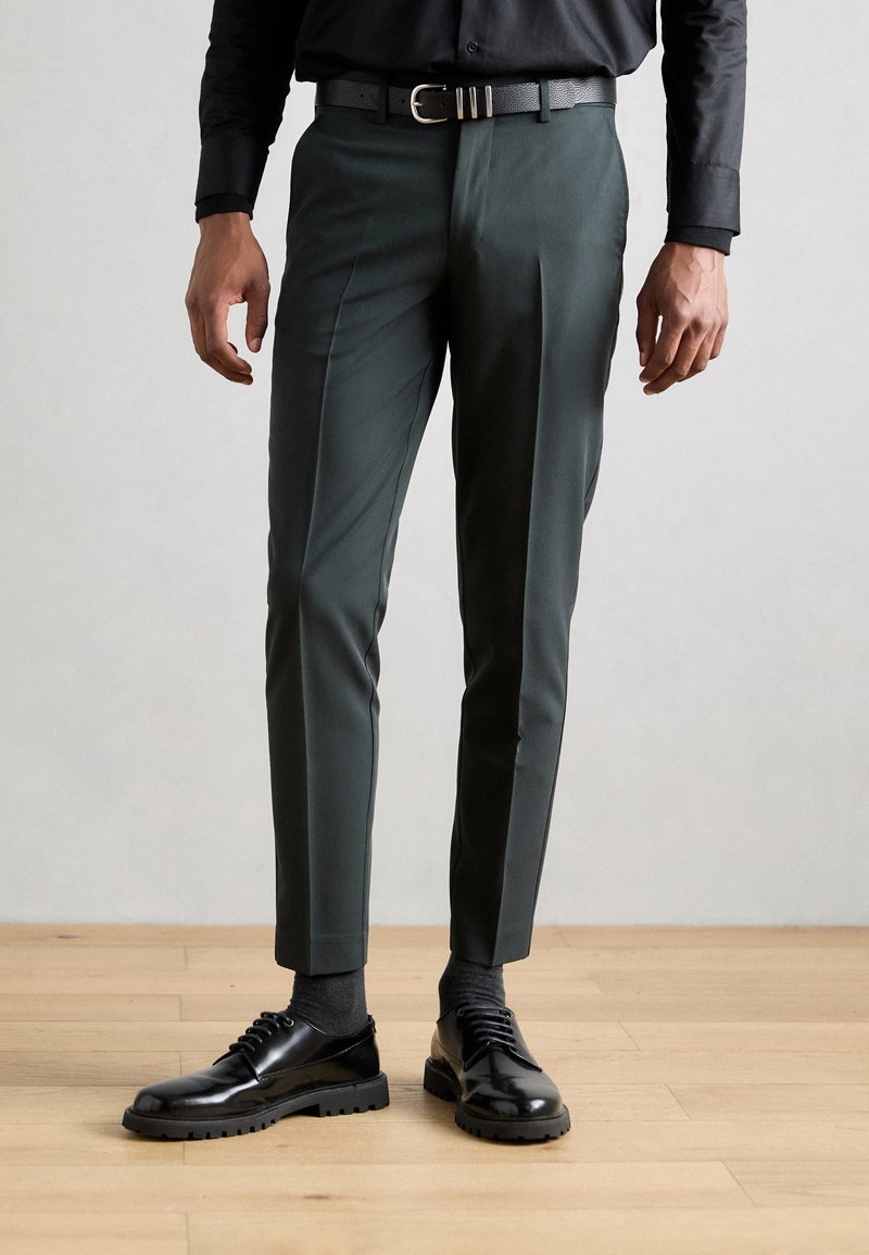 Úzké tmavě zelené kalhoty s plisováním, doplněné černým opaskem a lesklými černými botami. Povrch je hladký s lesklým povrchem.