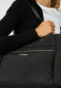 Schwarze Puffer-Tasche mit einer glatten, strukturierten Oberfläche, einem Reißverschluss-Frontfach und schwarzen Lederträgern. Logo auf der Tasche angebracht.