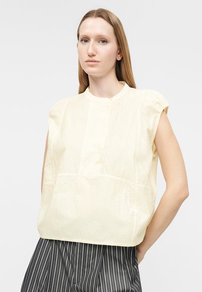 Γυναίκα με ίσια μαλλιά που φοράει αμάνικο, φαρδύ μπεζ πουκάμισο και μαύρο παντελόνι με λεπτές ρίγες, στέκεται μπροστά σε απλό ανοιχτόχρωμο φόντο.