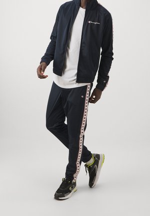 Homme portant un survêtement Champion bleu marine avec des bandes blanches et rouges du logo et des baskets noires avec des accents vert citron, debout sur fond blanc.