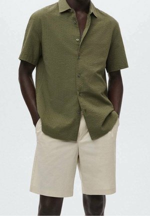 Homme portant une chemise boutonnée à manches courtes vert olive texturée avec un short beige longueur genou, les mains dans les poches, debout devant un fond uni.