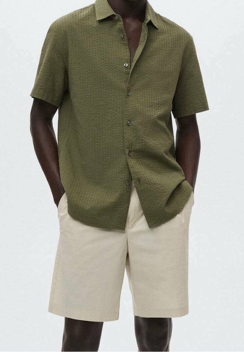 Homme portant une chemise boutonnée à manches courtes vert olive texturée avec un short beige longueur genou, les mains dans les poches, debout devant un fond uni.