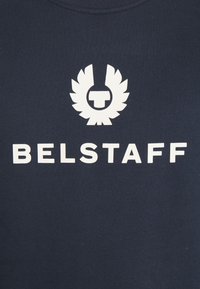 Vitt Belstaff-logotyp och namn tryckt på en mörk marinblå tygbakgrund, med en stiliserad vingemblem ovanför texten.