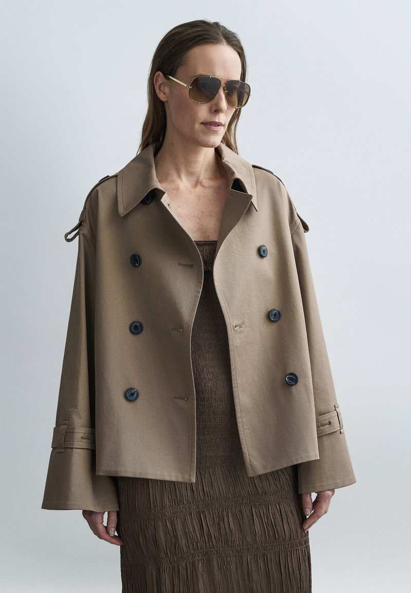 Bruine double-breasted trenchcoat met grote zwarte knopen, brede mouwen en schouderdetails. Draagt over een getextureerde bruine jurk.