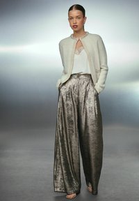 Dicker Strick-Cardigan in Creme mit einem mit Juwelen besetzten Ausschnitt, kombiniert mit weit geschnittenen metallisch-silbernen Hosen und einem weißem Top mit Spitze.