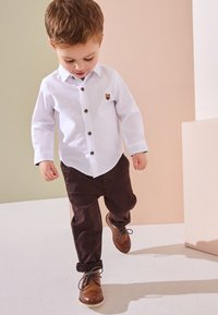 Camicia bianca a maniche lunghe con bottoni marroni e ricamo di un orso, abbinata a pantaloni marroni e scarpe marroni, mentre cammina su uno sfondo chiaro.