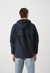 Giacca leggera blu navy con cappuccio, dotata di yoke posteriore, polsini elasticizzati e una silhouette rilassata per maggiore libertà di movimento.