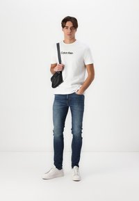 Camiseta blanca de algodón con el logo "Calvin Klein", jeans de mezclilla azules y zapatillas blancas. Bolso cruzado negro llevado sobre el hombro. Sencillo y casual.