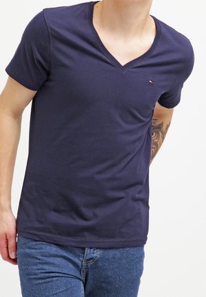 Person iført en mørkeblå V-hals t-shirt og blå jeans, med en synlig tatovering på højre underarm mod en ensfarvet baggrund.