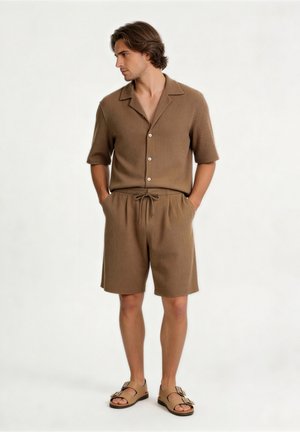 Mann trägt passendes braunes kurzärmliges Hemd mit Knöpfen und Shorts mit Kordelzug sowie hellbraune Sandalen mit Doppelriemen, steht mit den Händen in den Taschen.