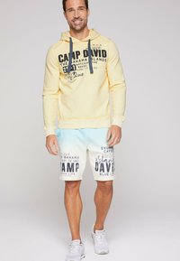 Sudadera amarilla con cordón, que presenta texto impreso en negro. Pantalones cortos azul claro y blancos con diseños gráficos, combinados con zapatillas blancas.