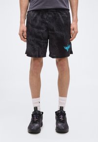 Άνδρας που φοράει μαύρο αθλητικό σορτς με υφή και μπλε λογότυπο φοίνικα, μαύρα αθλητικά παπούτσια και λευκές κάλτσες, στέκεται μπροστά σε απλό φόντο.