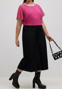 Haut en chiffon fuchsia avec un décolleté orné de bijoux, associé à un pantalon noir fluide et des bottes à plateforme noires montant jusqu'aux genoux. Petit sac à main noir avec des pierres précieuses.