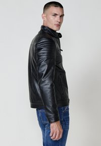 Giubbotto in pelle nera con dettagli a coste sulle spalle, chiusura con zip e tasche laterali. Indossato sopra jeans blu, presenta una silhouette aderente.