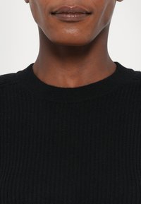 Zwarte geribbelde trui met een ronde halslijn, met een soepele textuur en subtiele details op de schouders.