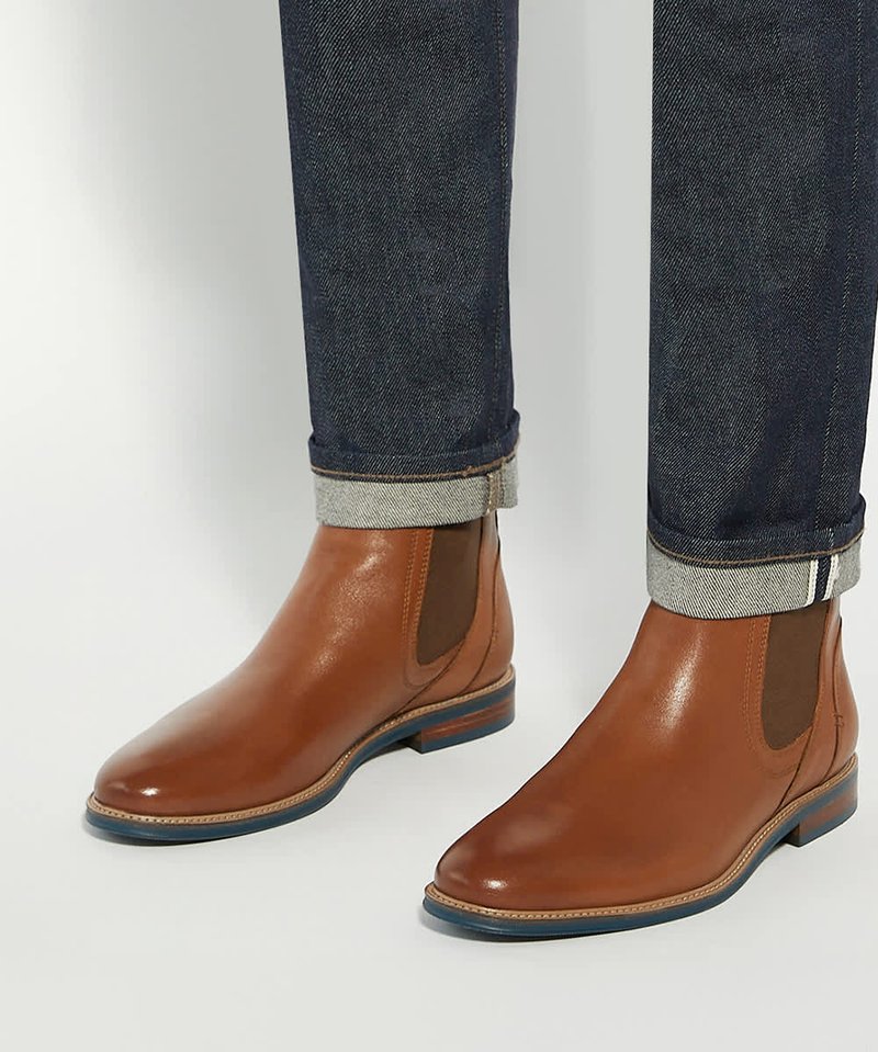 Paire de bottines Chelsea en cuir marron portées avec un jean bleu foncé retroussé sur un fond uni.