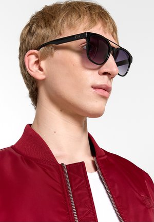 Giovane con capelli corti castano chiaro che indossa occhiali da sole neri e una giacca rossa con cerniera sopra una camicia bianca, guardando leggermente verso l'alto e a destra.
