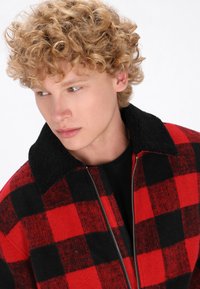 Rot- und schwarz kariertes Zip-Jackett mit einem schwarzen Fleece-Kragen, aus einem strukturierten Wollmischgewebe und mit einem kontrastierenden dunklen Hintergrund.