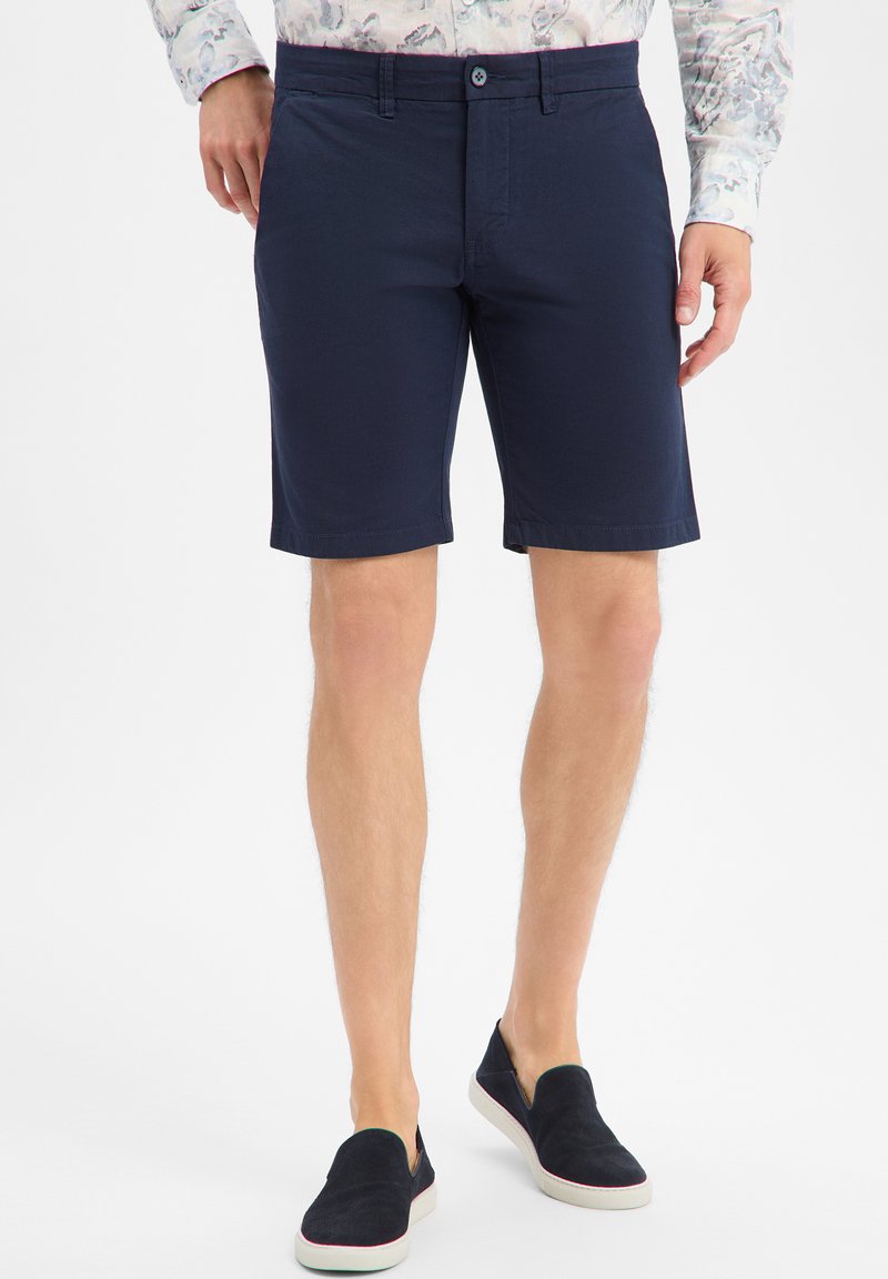 Man draagt marineblauwe tailored shorts, een bloemenprint blouse met lange mouwen en zwarte slip-on schoenen met witte zolen, staand op een witte achtergrond.