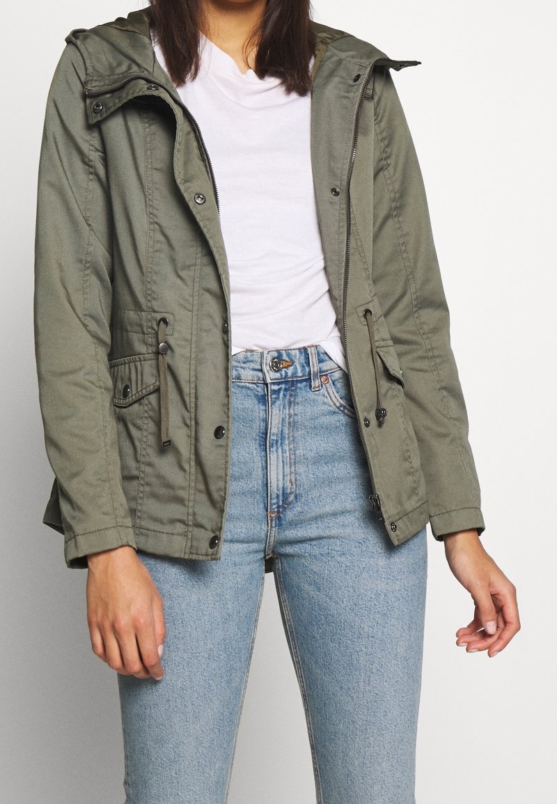 Kvinna som bär en olivgrön zip-up jacka över en vit skjorta och ljusblå högmidjade jeans, står mot en enfärgad bakgrund.