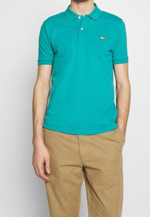 Mann trägt türkisfarbenes Poloshirt mit kurzen Ärmeln und gesticktem Logo sowie beigefarbene Hose, steht mit einer Hand in der Tasche.