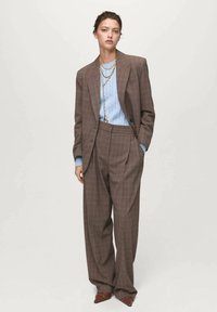 Brown plaid suit with wide-leg trousers and a matching oversized blazer over a light blue cable-knit sweater; layered gold chains as accents.