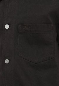 Poche de veste en denim noir avec un subtil logo de crocodile noir brodé et deux boutons en métal argenté visibles à l'avant.