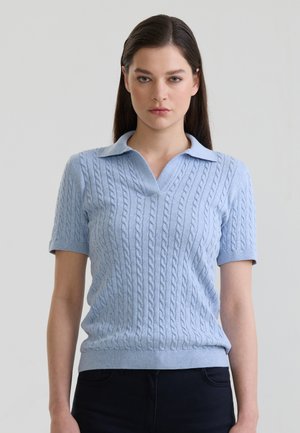Frau mit glatten dunklen Haaren trägt einen hellblauen kurzärmeligen Kabelstrick-Polo-Pullover und dunkle Hosen vor einem einfarbigen Hintergrund.