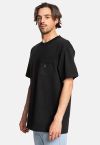 Černé tričko s krátkým rukávem, které má na přední straně kapsu s logem, vyrobené z bavlny, s uvolněným střihem a jednolitou strukturou.