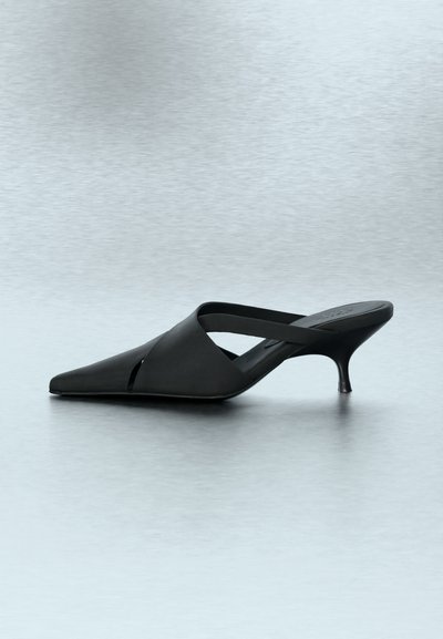 Sort mule med spids tå, mellemhøj kittenhæl og en diagonal rem over foden på en grå overflade.