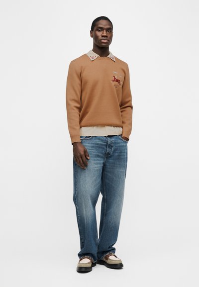 Άνδρας που στέκεται φορώντας πλεκτό πουλόβερ σε καφέ απόχρωση με κέντημα αλόγου, μπεζ πουκάμισο με γιακά από κάτω, άνετο μπλε τζιν και μπεζ και καφέ μοκασίνια.