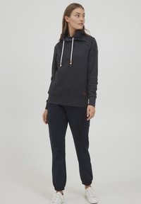 Felpa grigia scura con colletto alto, cordini bianchi e bottoni di legno, abbinata a pantaloni jogger blu navy con tasche laterali.