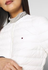 Femme portant une veste blanche matelassée sur un col roulé gris clair, avec un petit logo rouge, blanc et bleu sur la poitrine de la veste.