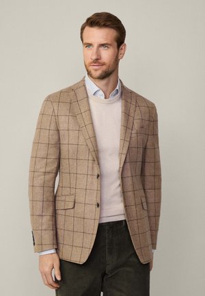 Brauner karierter Blazer aus strukturiertem Stoff, mit zwei Knöpfen, Reverskragen und Fronttaschen, über einem hellen Pullover getragen.