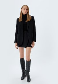 Zwarte op maat gemaakte blazer over een zwarte geribbelde turtleneck, gepaard met een zwarte minirok en zwarte kniehoge laarzen met hakken.