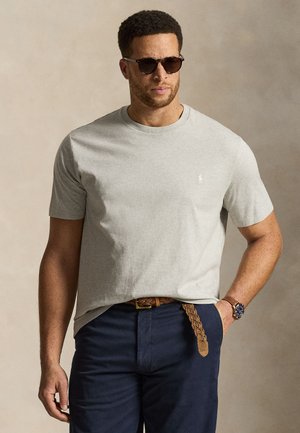 Férfi szürke rövid ujjú pólóban, sötétkék nadrágban, fonott barna övvel, órával és napszemüveggel, áll egy kézzel a zsebében.