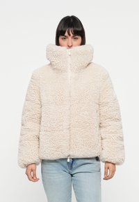 Beige nepbontjas met een hoge kraag, rits aan de voorkant en elastische zoom. Het materiaal is gestructureerd en pluizig, wat warmte biedt.
