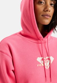 Mujer vistiendo una sudadera rosa brillante con un logo en forma de corazón y el texto "roxy", mostrada de los hombros hacia arriba contra un fondo liso.