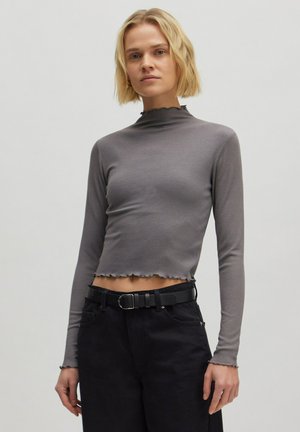 Grauer, gerippter, langärmeliger Cropped-Top mit hohem Kragen und Rüschensaum, kombiniert mit hoch taillierten schwarzen Hosen und einem schwarzen Gürtel.