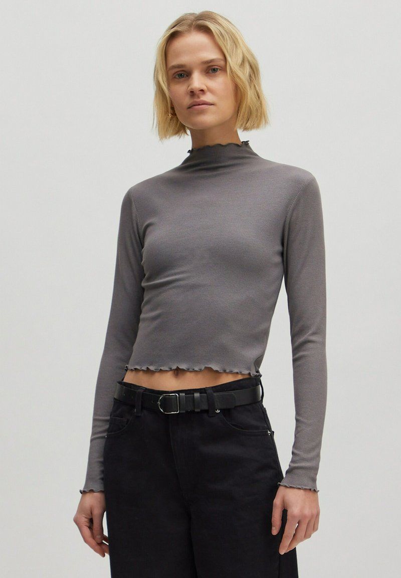 Grauer, gerippter, langärmeliger Cropped-Top mit hohem Kragen und Rüschensaum, kombiniert mit hoch taillierten schwarzen Hosen und einem schwarzen Gürtel.