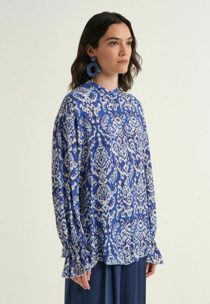 Donna che indossa una camicetta a maniche lunghe con motivo blu e bianco, con polsini arricciati e grandi orecchini circolari blu, in piedi davanti a uno sfondo semplice.