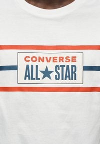 Λευκό βαμβακερό μπλουζάκι με τολμηρό λογότυπο "CONVERSE ALL STAR" σε κόκκινο και μπλε, περιβαλλόμενο από δύο οριζόντιες ρίγες σε αντίθετα χρώματα.