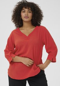 Blouse rouge texturée avec un décolleté en V, des manches trois-quarts et une coupe ample. L'ourlet est arrondi et le tissu semble léger.