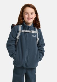 Giacca in fleece blu con zip, indossata sopra una camicia grigia. Presenta spallacci grigi regolabili, con un logo sull'area del petto a sinistra.