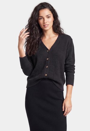 Ung kvinde med langt krøllet hår iført en sort knaplukket cardigan og sort ribbet nederdel, stående mod en ensfarvet lys baggrund.