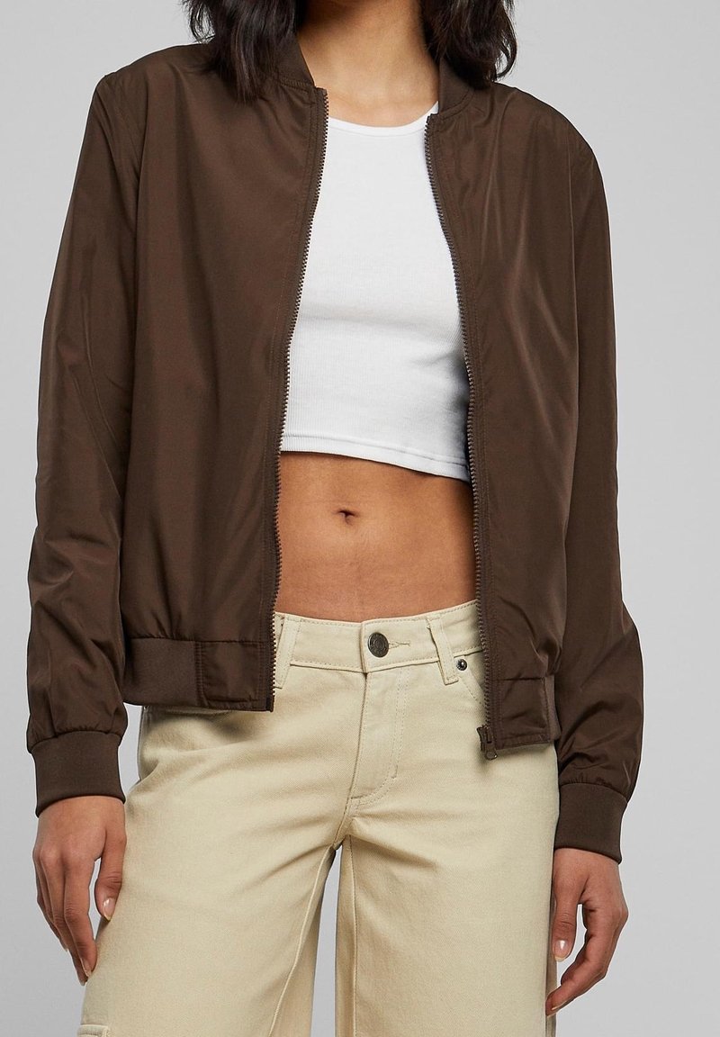 Chaqueta bomber marrón con cierre frontal, puños y cinturilla de canalé. Llevada sobre una camiseta corta blanca, combinada con pantalones de color claro. Textura suave.